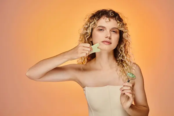 Charming curly woman with jade roller and gua sha looking at camera on pink and yellow backdrop — Stock Photo