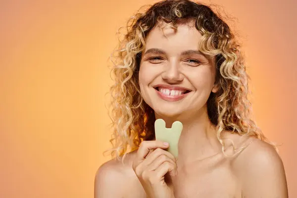 Cheerful curly woman with perfect skin holding gua sha looking at camera on pastel backdrop — Stock Photo