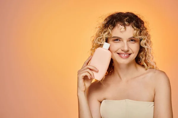 Sensual woman with wavy hair and glowing skin holding bottle of body lotion on pink and yellow — Stock Photo
