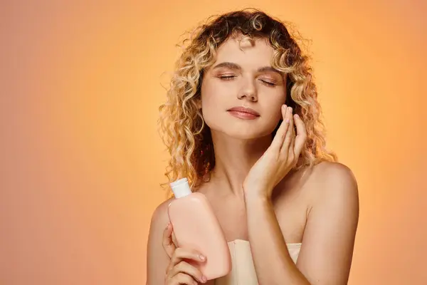 Delighted woman with wavy hair and perfect skin applying body lotion on pink and yellow backdrop — Stock Photo