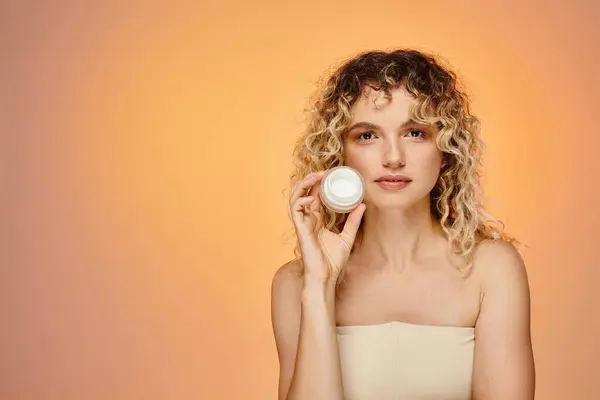 Attractive woman with wavy hair and natural makeup showing jar of face cream on yellow and pink — Stock Photo