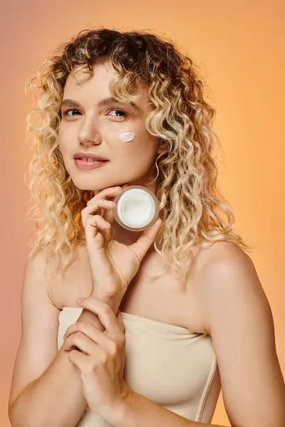Donna sorridente con i capelli ondulati e la pelle perfetta che mostra vaso di crema viso su sfondo pastello — Foto stock