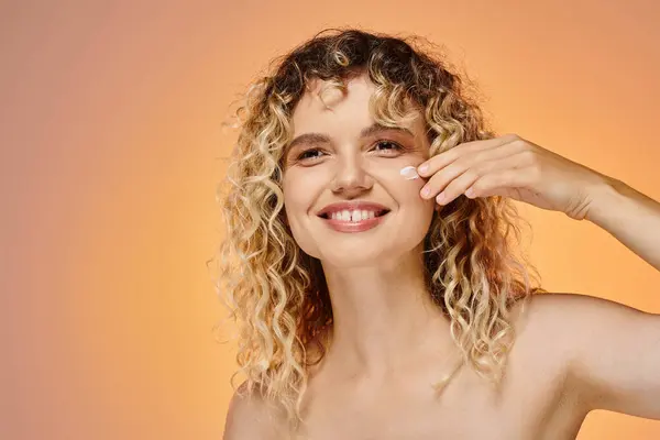 Щаслива жінка з хвилястим волоссям наносить косметичний крем на ідеальне обличчя на жовтий і рожевий фон — Stock Photo