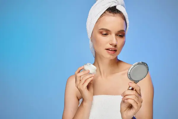 Mulher encantadora com creme facial e toalha na cabeça olhando para o espelho no fundo azul, auto-cuidado — Fotografia de Stock