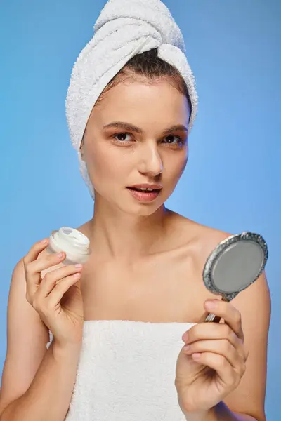 Femme attrayante avec crème pour le visage et serviette sur la tête en regardant la caméra sur fond bleu, auto-soins — Photo de stock