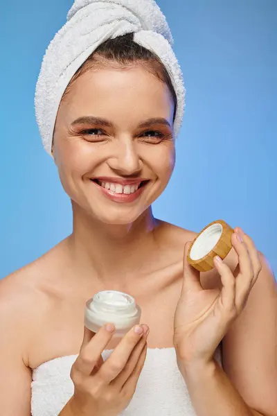 Femme heureuse avec serviette sur la tête tenant pot de crème pour le visage et regardant caméra sur bleu — Photo de stock