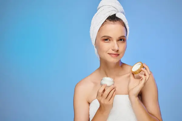 Sensual woman with towel on head holding jar of face cream and looking at camera on blue — Stock Photo