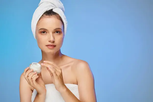 Sensual bonita mujer con toalla en la cabeza celebración de crema facial una mirada a la cámara en el fondo azul - foto de stock