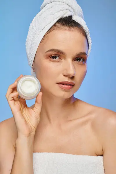 Atractiva mujer con toalla en la cabeza mostrando tarro de crema facial sobre fondo azul, bienestar y belleza - foto de stock