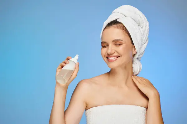 Cheerful woman with towel on head and perfect face holding dispenser with cosmetic foam on blue — Stock Photo
