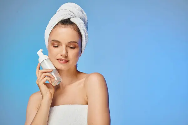 Mulher sorridente com pele brilhante e toalha na cabeça segurando dispensador com espuma facial no azul — Fotografia de Stock