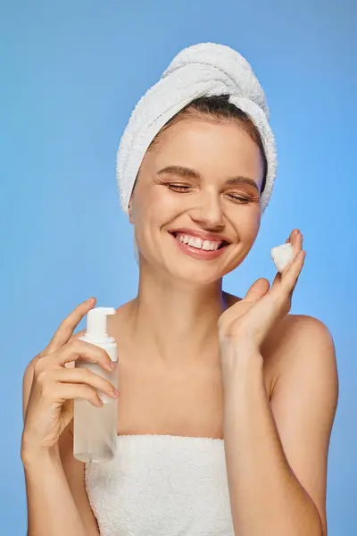 Delighted woman with closed eyes and towel on head holding bottle of face foam on blue backdrop — Stock Photo