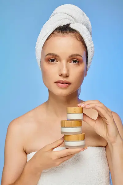 Séduisante femme avec peau propre et serviette sur la tête tenant crème cosmétique regardant caméra sur bleu — Photo de stock