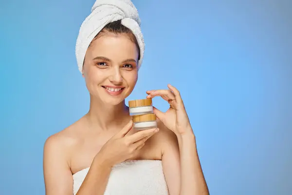 Delighted woman with perfect skin and towel on head holding cosmetic cream looking at camera on blue — Stock Photo