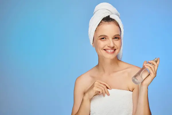 Cheerful woman with towel on head and bottle of body spray looking at camera on blue backdrop — Stock Photo