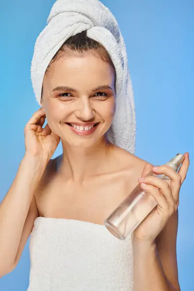 Femme heureuse avec serviette sur la tête et bouteille de spray corporel souriant à la caméra sur fond bleu — Photo de stock