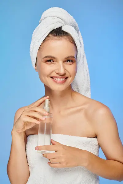 Mulher feliz com toalha na cabeça e garrafa de spray corporal sorrindo para a câmera no fundo azul — Fotografia de Stock