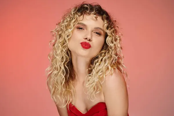 Flirty curly woman with bold makeup pouting lips while sending air kiss at camera on pastel backdrop — Stock Photo