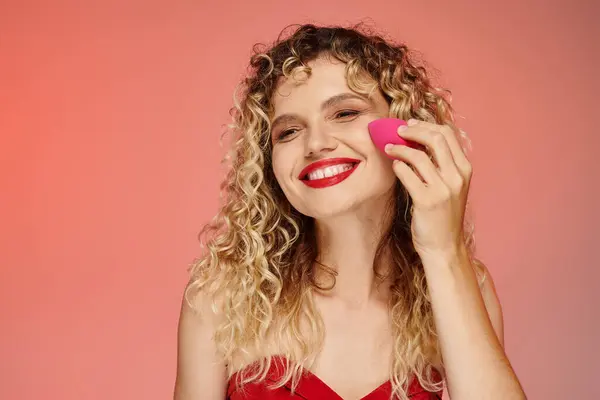 Curly cheerful woman with red lips using face sponge on pink and yellow backdrop, elegant beauty — Stock Photo