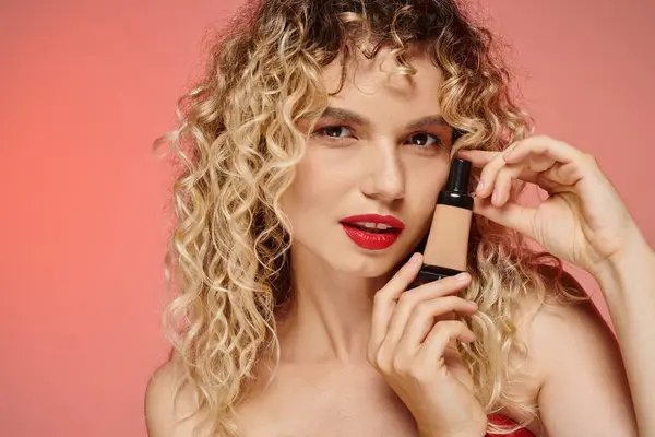 Faszinierende Frau mit roten Lippen und welligem Haar zeigt Make-up Foundation auf pastellrosa Hintergrund — Stockfoto