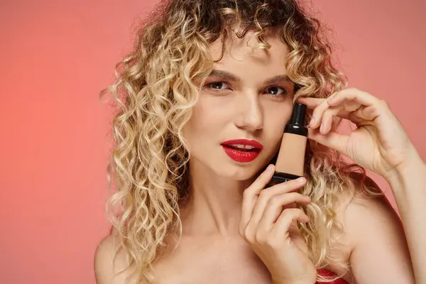 Mesmerizing woman with red lips and wavy hair showing makeup foundation on pastel pink backdrop — Stock Photo