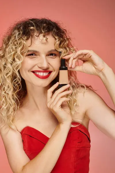 Cheerful pretty woman with curly hair and perfect skin showing makeup foundation on pink backdrop — Stock Photo