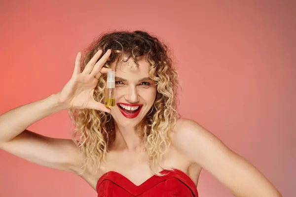 Overjoyed woman with curly hair and red lips holding makeup foundation on pink and yellow backdrop — Stock Photo