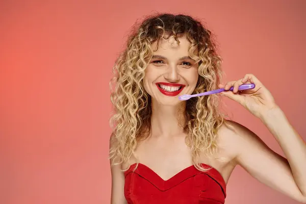 Fröhliche lockige Frau mit roten Lippen und Zahnbürste, die vor pastellfarbenem Hintergrund in die Kamera blickt — Stockfoto