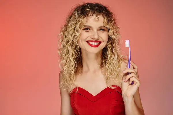 Faszinierende Frau mit welligem Haar und roten Lippen, die eine Zahnbürste in der Hand hält und in die Kamera schaut — Stockfoto