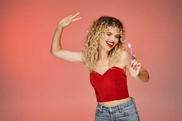 Excited woman with wavy hair and red lips dancing with toothbrush on pink and yellow backdrop — Stock Photo
