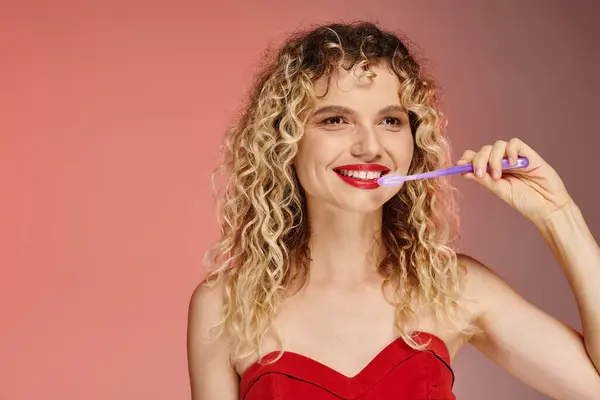 Attractive curly woman with red lips and radiant smile cleaning teeth on pastel pink backdrop — Stock Photo