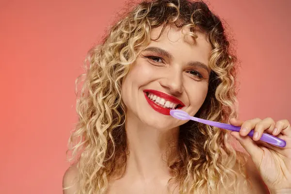 Encantadora mujer rizada con labios rojos y sonrisa radiante limpieza de dientes sobre fondo rosa pastel - foto de stock