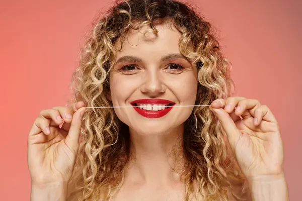 Porträt einer fröhlichen Frau mit welligem Haar und roten Lippen, die Zähne mit Zahnseide auf rosa putzt — Stockfoto