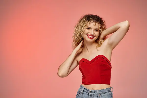 Woman with red lips and curly hair posing in red top and smiling at camera on pastel backdrop — Stock Photo