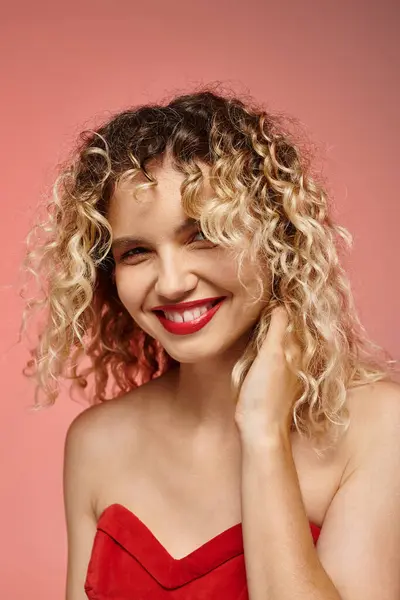 Retrato de mujer excitada con el pelo rizado y la parte superior roja sonriendo a la cámara en el fondo de color rosa pastel - foto de stock