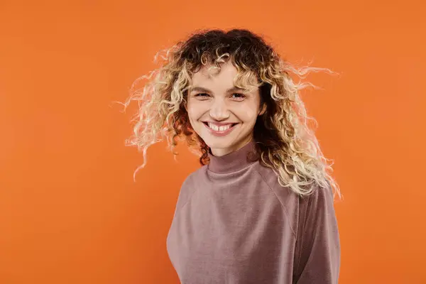 Cheerful woman with wavy hair in mocha color turtleneck looking at camera on radiant orange backdrop — Stock Photo