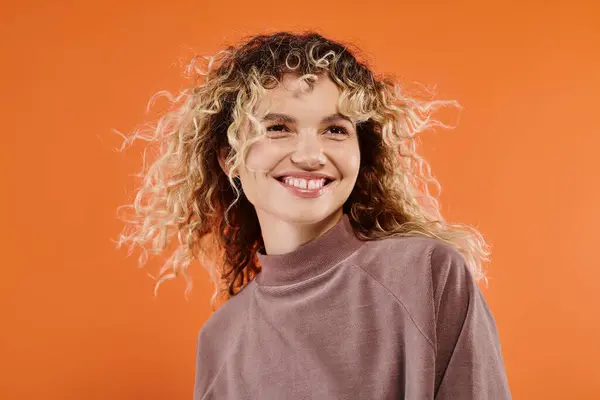 Carefree woman with curly hair in mocha color turtleneck looking at camera on bright orange backdrop — Stock Photo