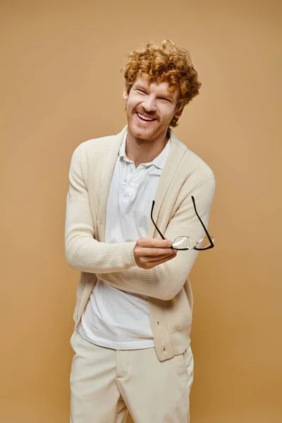 Hombre pelirrojo excitado en ropa de color claro sosteniendo anteojos y riendo sobre fondo beige - foto de stock