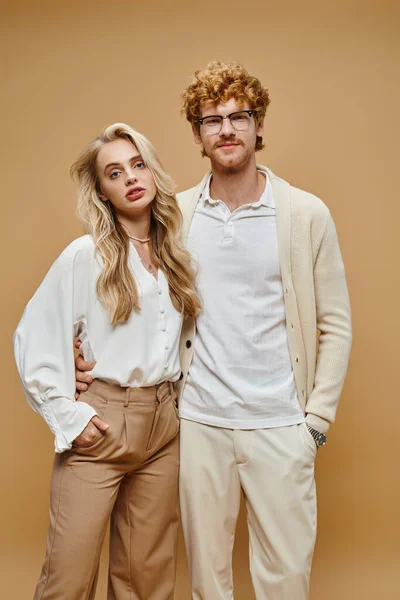 Trendy young couple in light-colored clothes with hands in pockets looking at camera on beige — Stock Photo