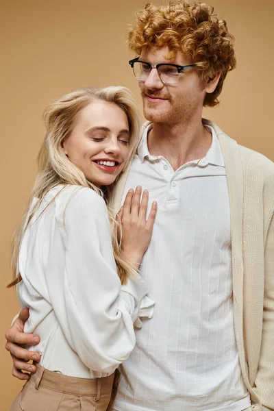 Glücklicher rothaariger Mann mit Brille umarmt stylische blonde Frau lächelnd mit geschlossenen Augen auf beige — Stockfoto