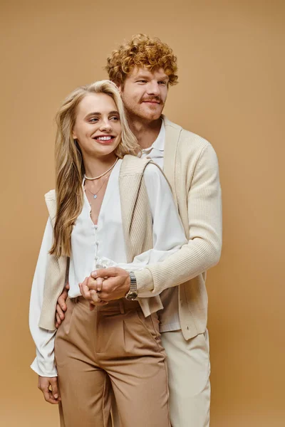 Positive blonde woman and redhead man in light clothes holding hands and looking away on beige — Stock Photo
