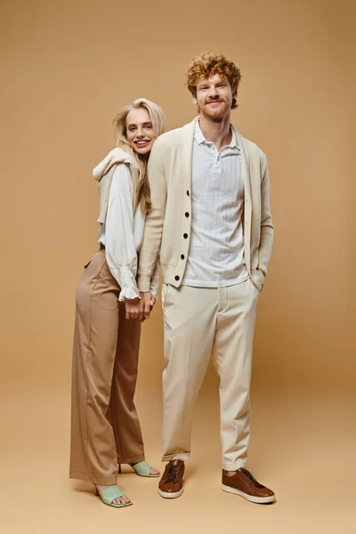 Full length of happy young couple in trendy casual clothes holding hands looking at camera on beige — Stock Photo