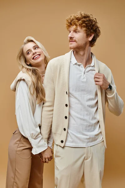 Old money style, young cheerful couple in trendy outfit holding hands and looking away on beige — Stock Photo