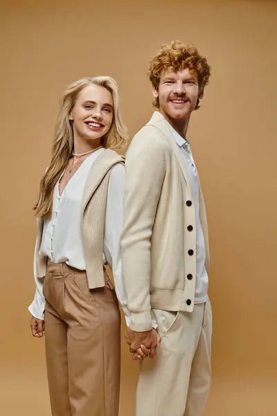 Fashionable joyous couple in trendy casual clothes standing back to back and holding hands on beige — Stock Photo