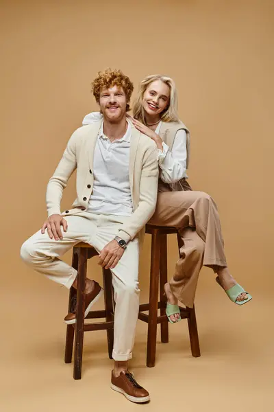 Alegre jovem casal em roupas elegantes sentado em cadeiras e olhando para a câmera no fundo bege — Fotografia de Stock
