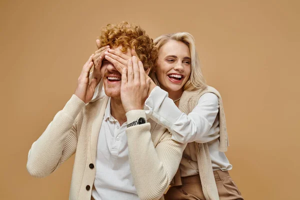 Playful blonde woman covering eyes of stylish redhead man on beige, old money style fashion — Stock Photo