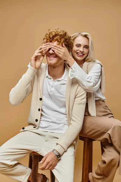 Mulher loira animado sentado na cadeira e cobrindo os olhos do homem ruiva na moda no fundo bege — Fotografia de Stock