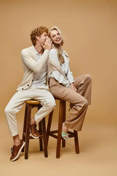 Fashionable redhead man telling secret to trendy blonde woman while sitting on chairs on beige — Stock Photo