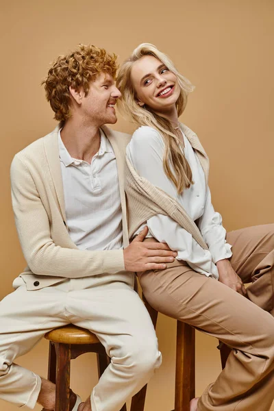 Jovem e sorrindo casal em trajes casuais na moda sentado em cadeiras em cinza, moda estilo dinheiro velho — Fotografia de Stock