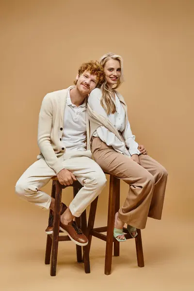 Comprimento total de feliz jovem casal em roupas de cor clara sentado em cadeiras em pano de fundo bege — Fotografia de Stock